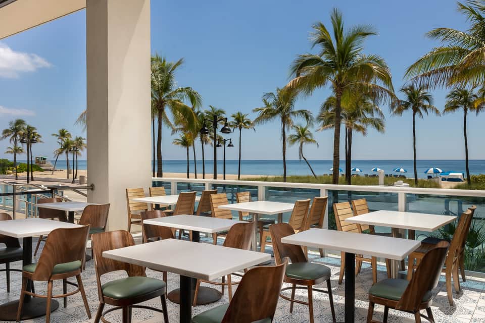 Beachfront dining area with wooden chairs, white tables, and ocean view beyond palm trees
