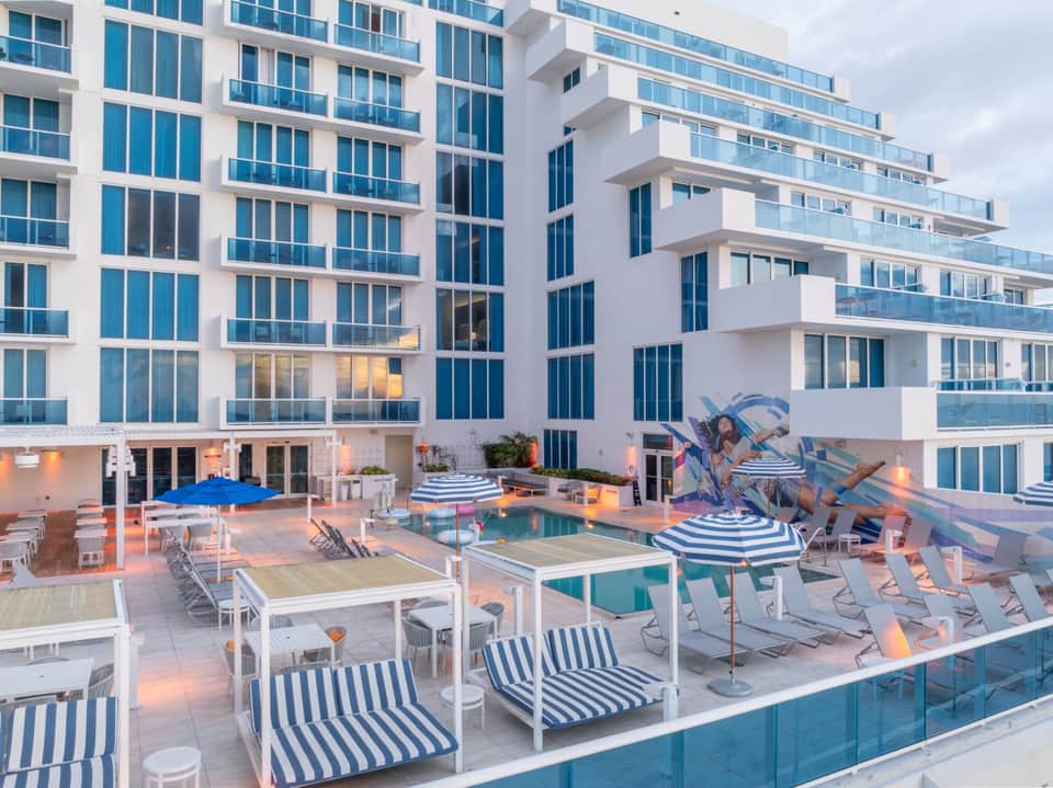 Modern white high-rise resort with multiple balconies, pool deck, and striped umbrellas at dusk