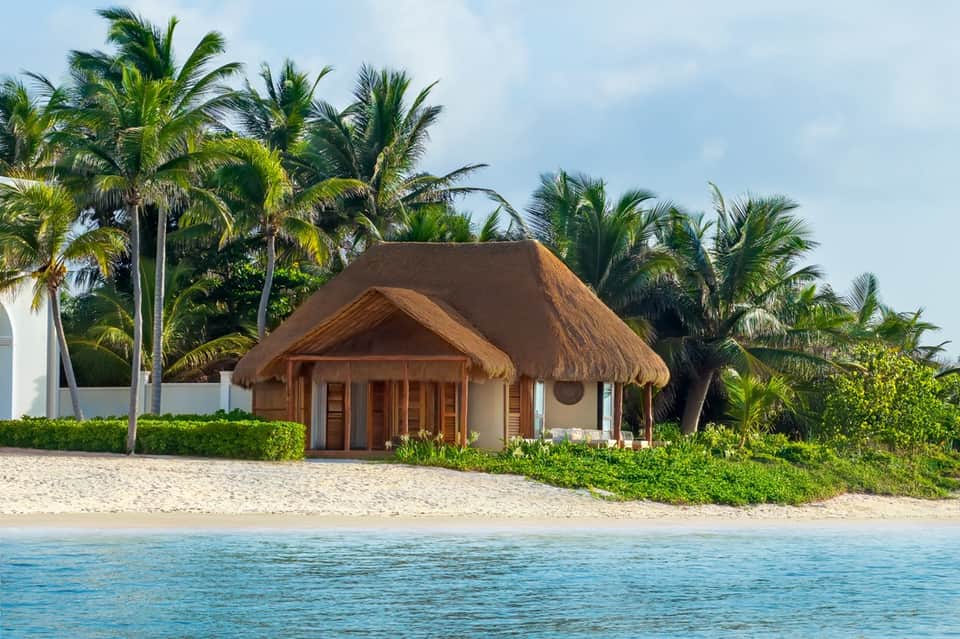 Beachfront thatched-roof bungalow with palm trees and white sand beach, tropical resort setting