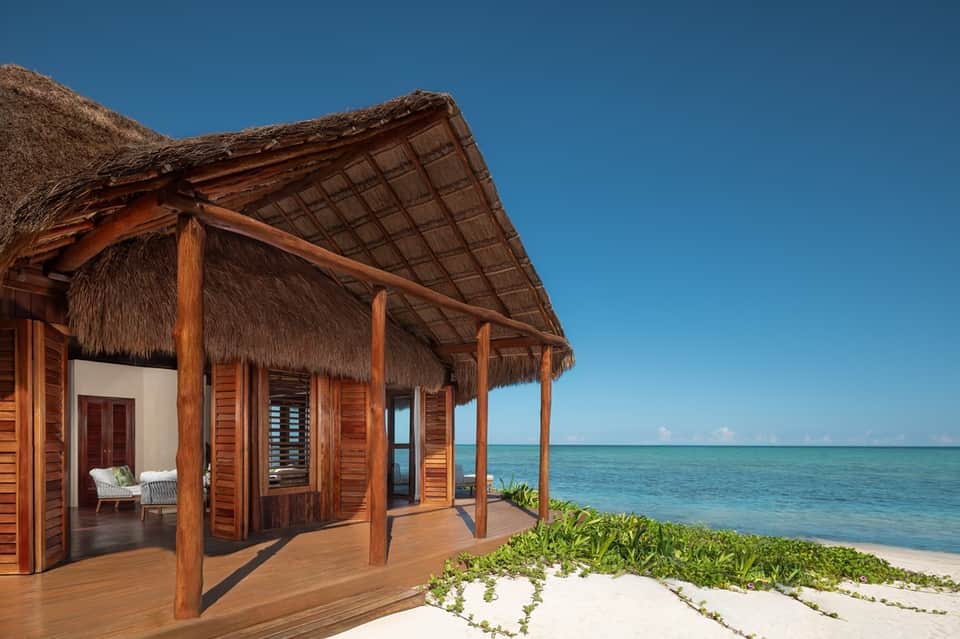 Beachfront wooden villa with thatched roof, covered deck, pristine white sand, and turquoise ocean