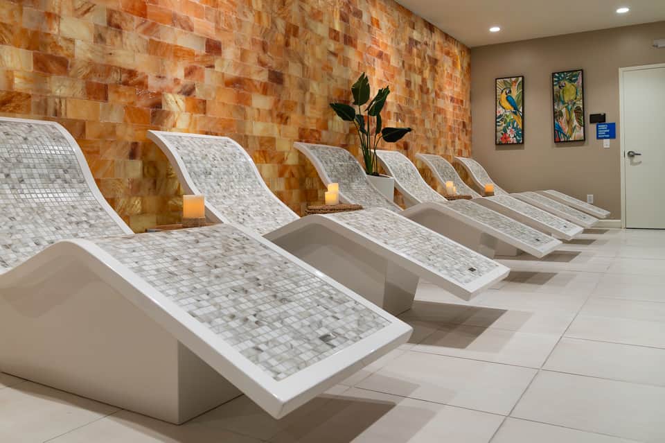 Modern spa relaxation area with multiple mosaic tile loungers, warm stone accent wall, decorative plants, and ambient lighting