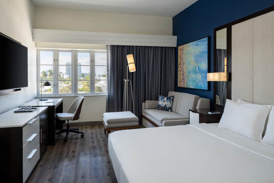 Modern hotel room with king bed, work desk, sitting area, dark curtains, and city skyline views through large windows