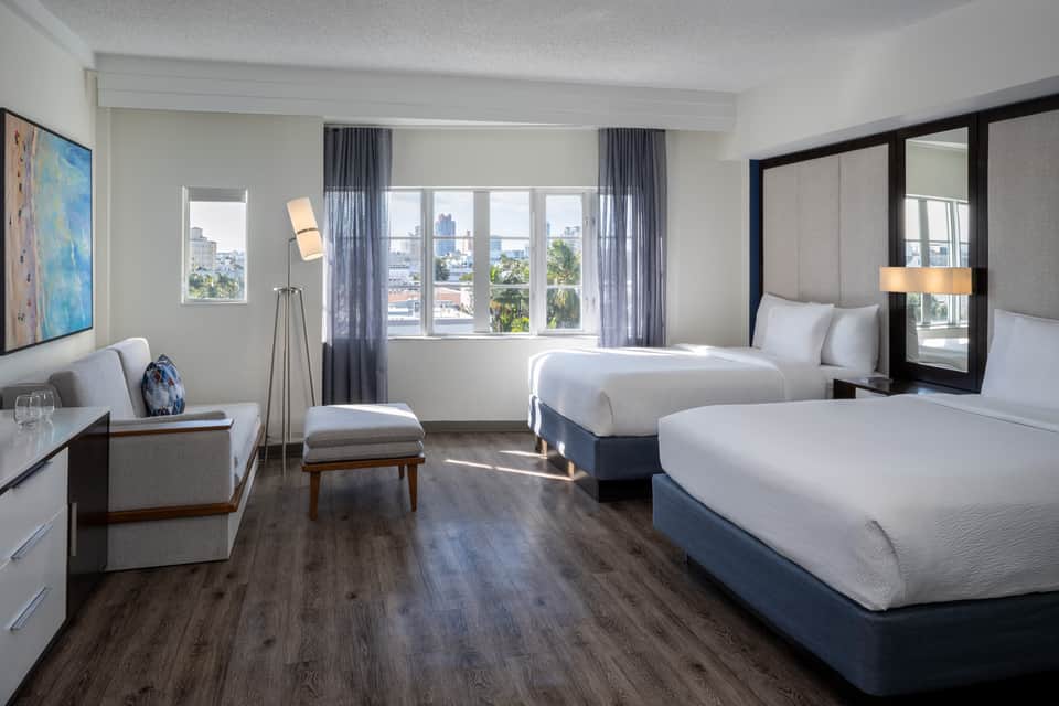Bright hotel room with two queen beds, lounge seating, desk area, and expansive windows overlooking cityscape