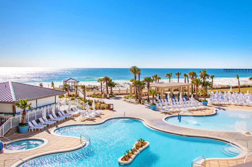 Beachfront resort pools with palm trees, lounge chairs, and ocean view with pier