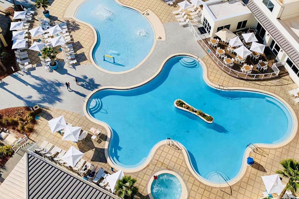 Aerial view of resort with multiple curved swimming pools, white umbrellas, and terracotta-tiled deck
