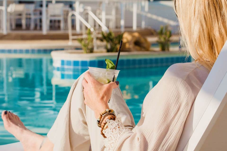 Woman holding a cocktail by the pool overlooking a resort with white buildings and palm trees