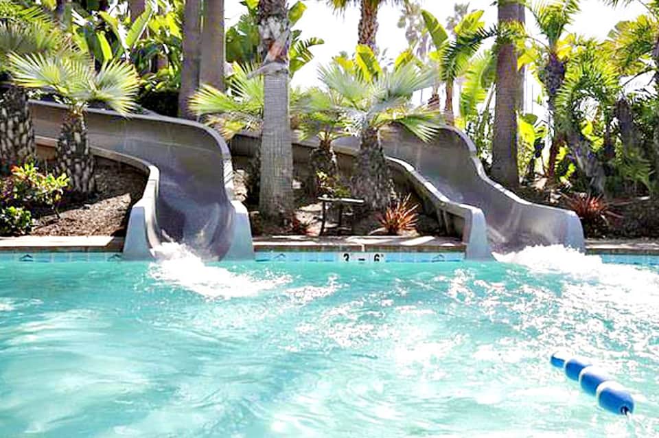 Resort pool with dual water slides surrounded by palm trees and tropical landscaping