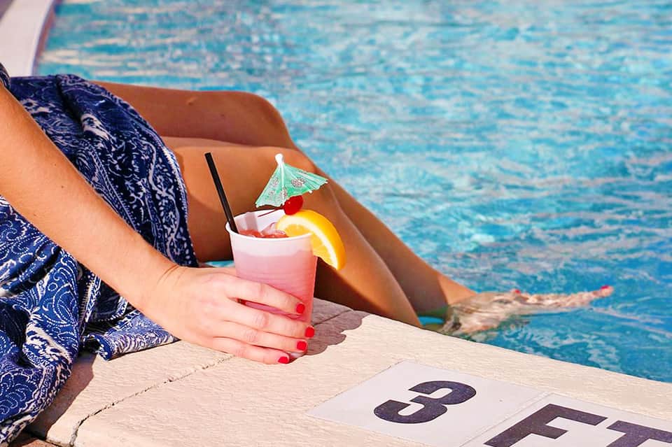 Woman holding pink cocktail with umbrella by pool edge in swimwear