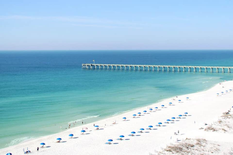 White sandy beach with blue umbrellas and a wooden pier extending into turquoise ocean