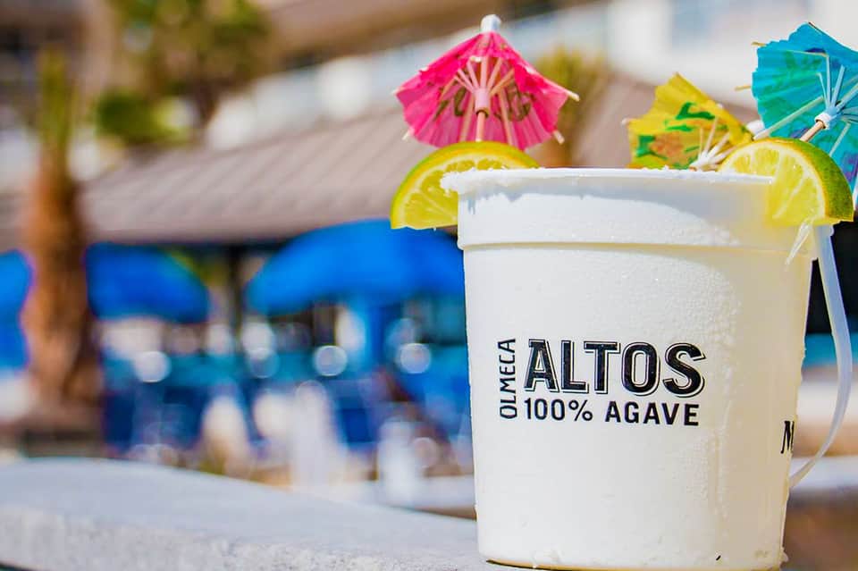 Frozen margarita drink with Altos 100% Agave label, lime slice, and colorful umbrellas by poolside