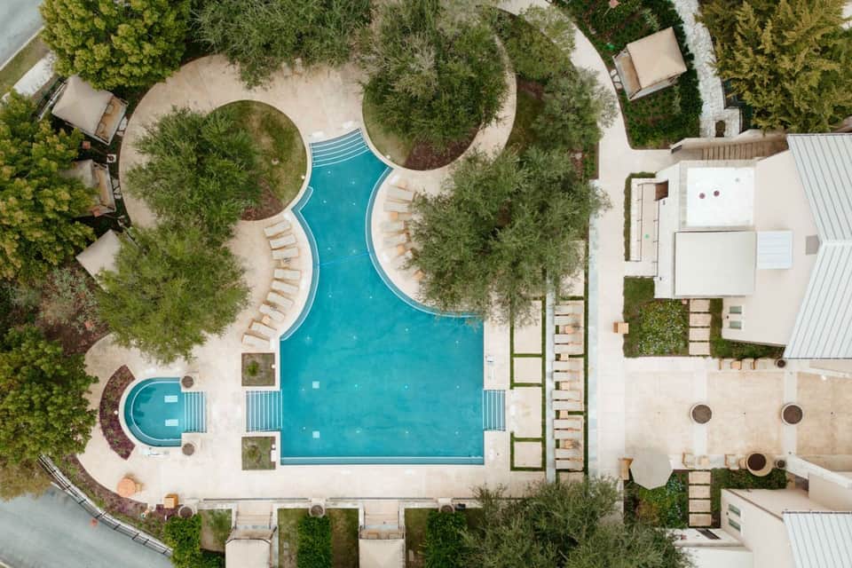 Aerial view of a curved resort pool surrounded by lounge chairs, manicured lawns, and cypress trees with building structures nearby