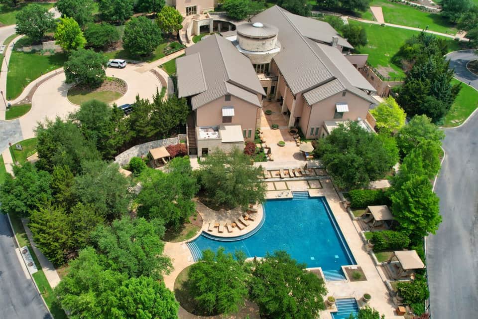 Aerial view of luxury resort with geometric roof design, curved pool with loungers, and manicured landscaping surrounded by trees