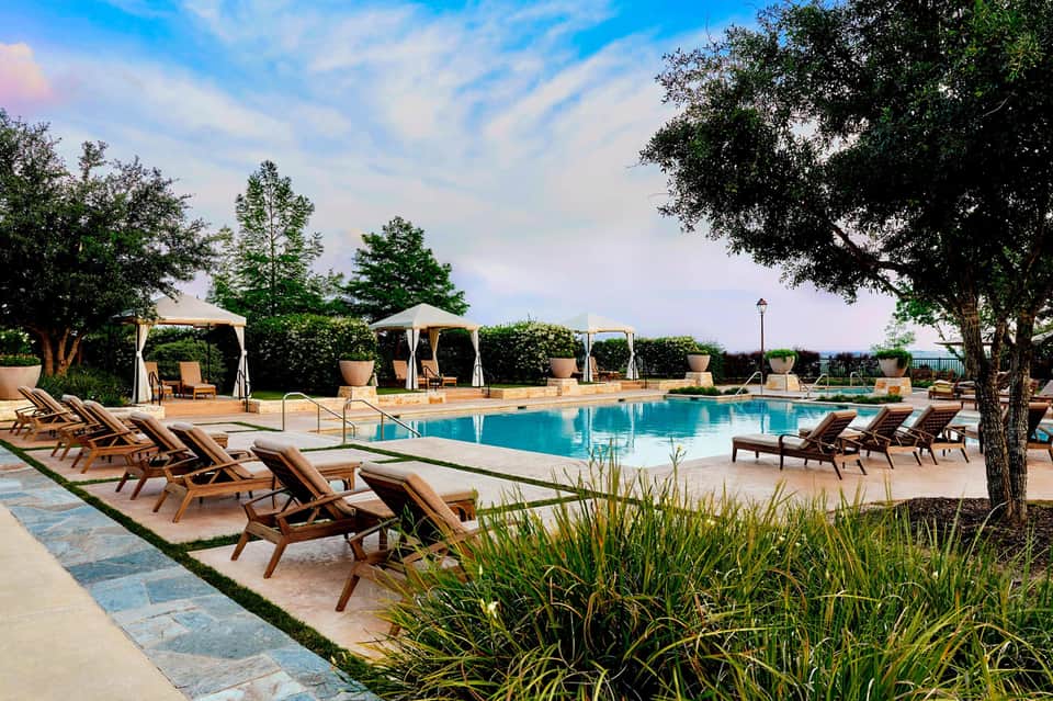 Resort pool with wooden lounge chairs, white cabanas, mature trees, and decorative landscaping