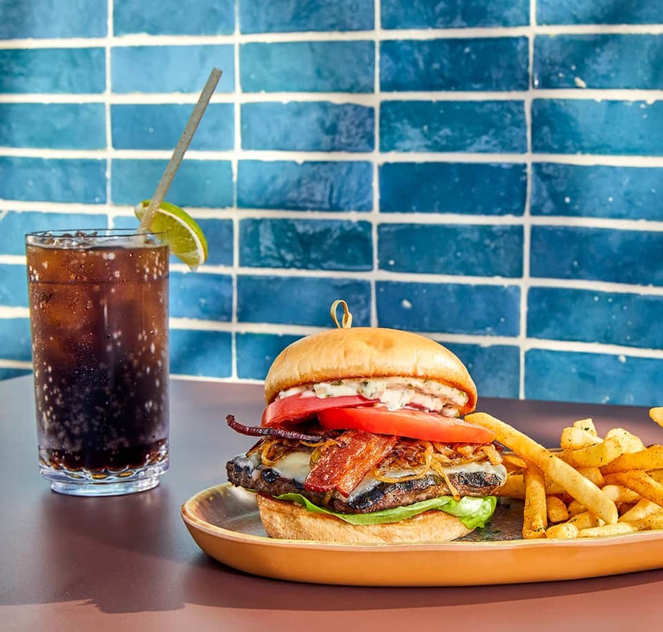 Gourmet burger with bacon and toppings, fries and cola drink against blue tile background