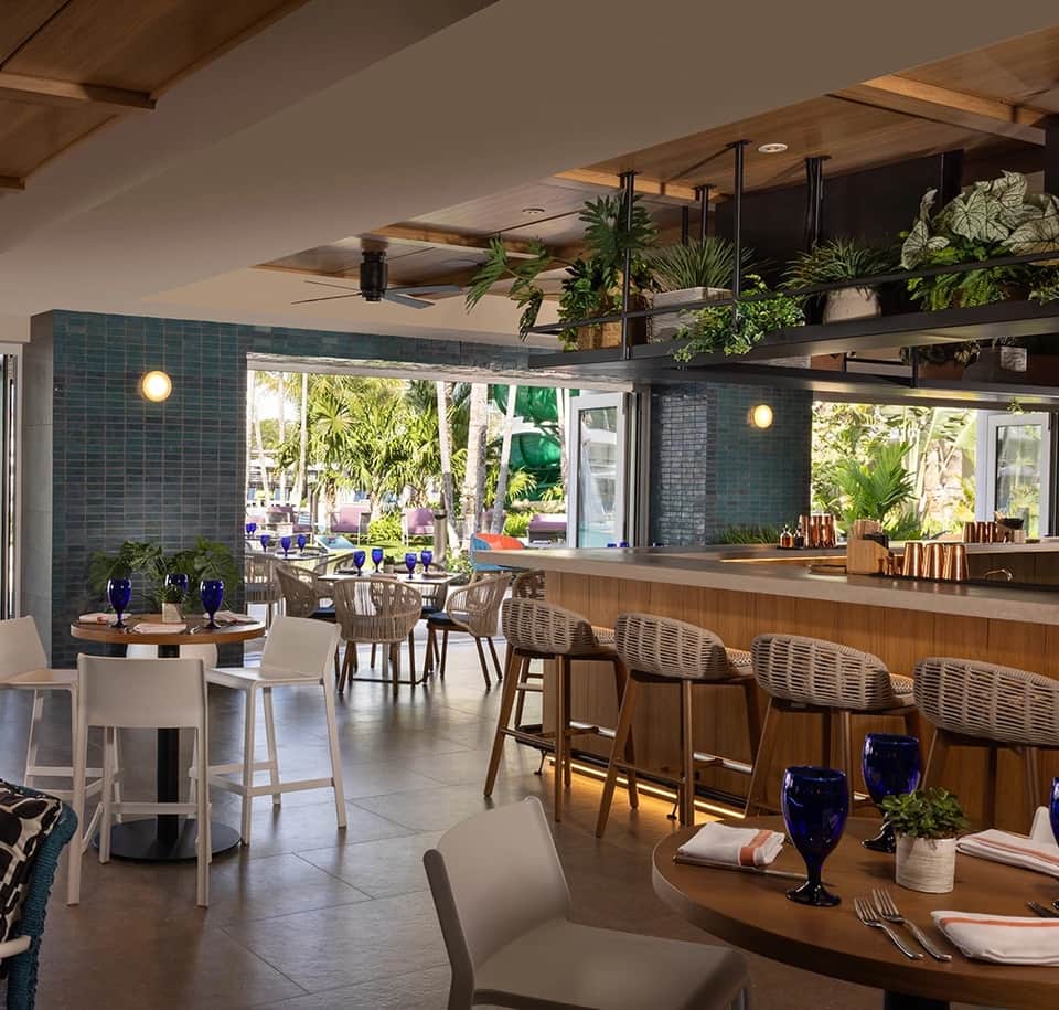Modern resort restaurant with blue tile walls, wooden bar seating, and tropical plants overlooking palm tree gardens
