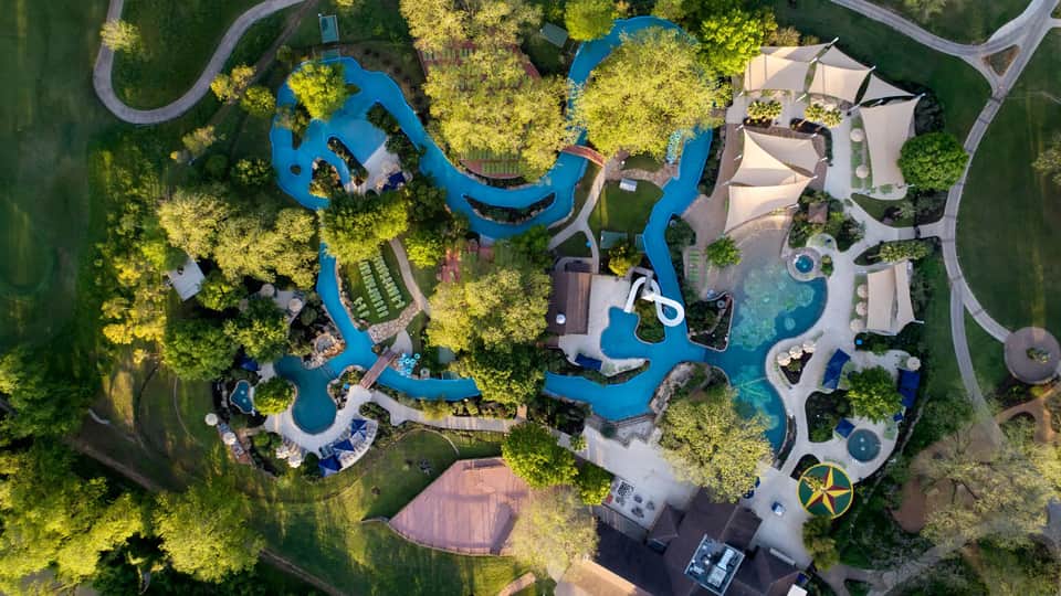 Aerial view of resort with interconnected blue water features, pools, landscaped grounds, and multiple buildings surrounded by green trees