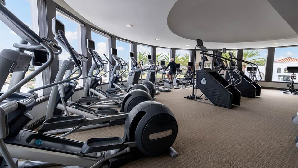 Modern fitness center with rows of elliptical machines and exercise equipment, floor-to-ceiling windows overlooking grounds
