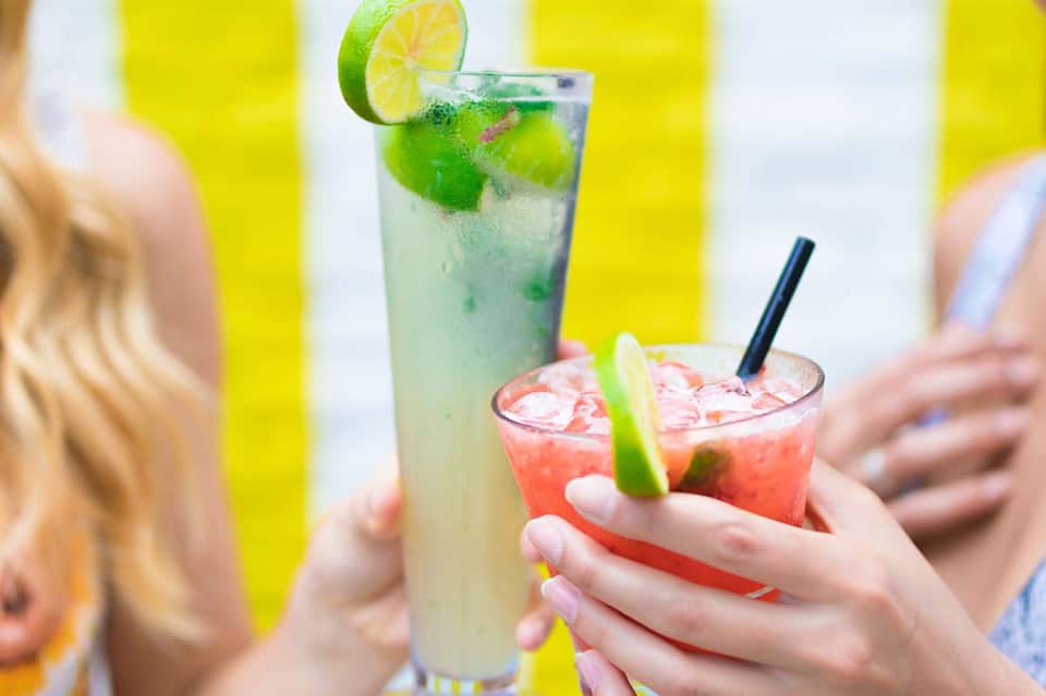 Colorful tropical cocktails with lime garnish held by hands against yellow striped background