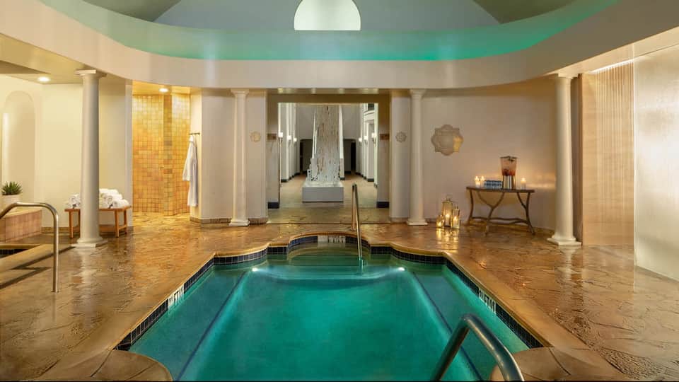 Luxurious indoor spa pool with turquoise water, columns, heated deck, and ambient lighting