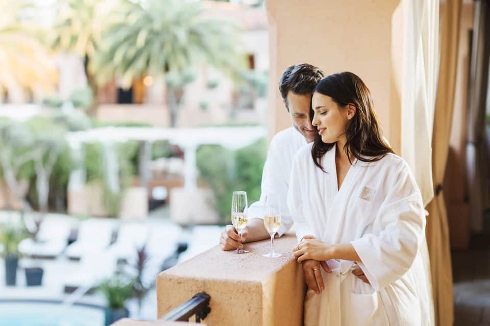 Couple in white robes enjoying champagne on terrace overlooking resort courtyard and gardens