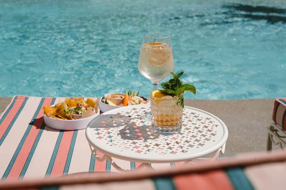 Poolside table with cocktail, appetizers, and pink menu on turquoise water background