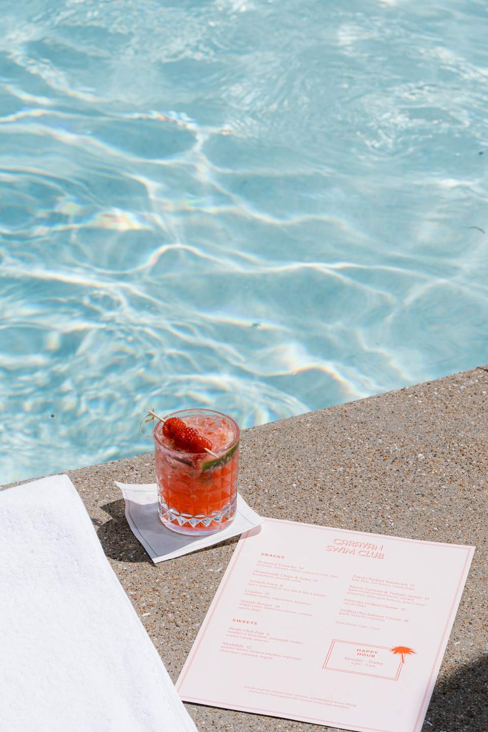 Red cocktail with strawberry on pool deck next to pink menu with turquoise water reflection