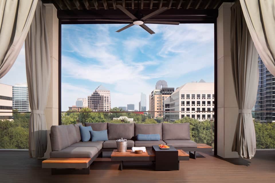 Modern suite terrace with sectional sofa, panoramic city views, curtains, and ceiling fan overlooking downtown