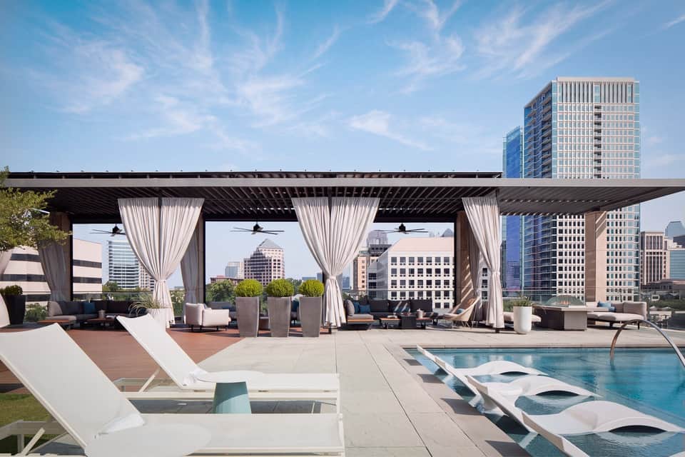 Rooftop lounge with pergola, curtains, lounge seating, infinity pool, and downtown skyline at daytime