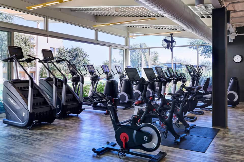 Fitness center with rows of cardio equipment, stationary bikes, and large windows overlooking trees