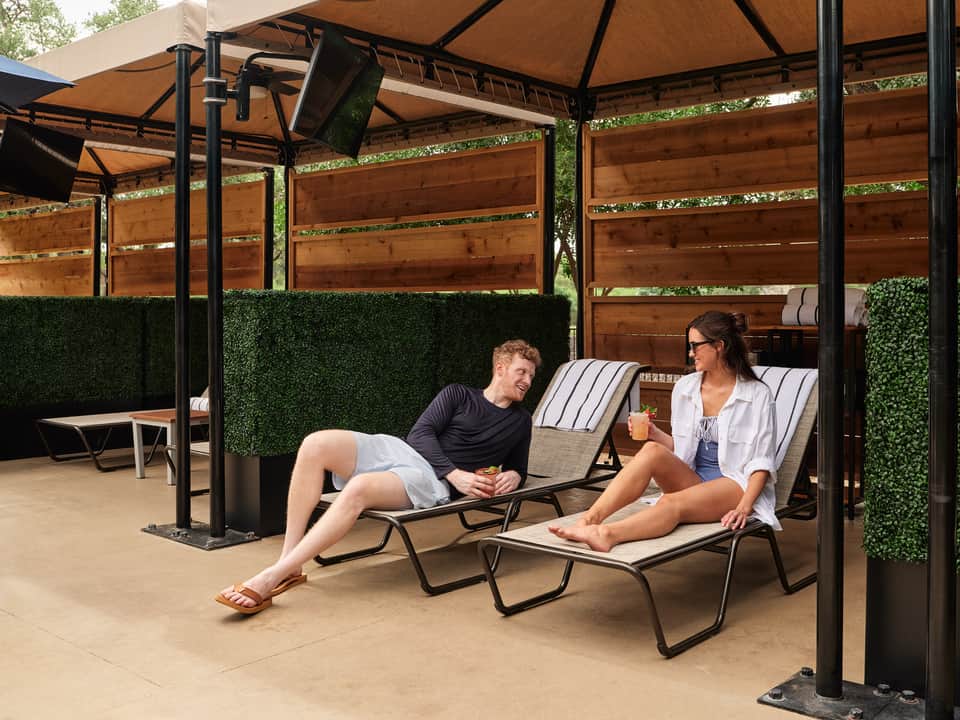 Two guests relaxing in lounge chairs under pergola with tan canopy and green artificial wall panels