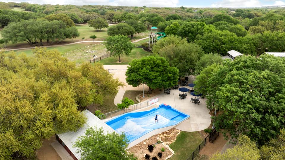 Aerial view of resort with blue wave-shaped pool, surrounding trees, playground, and sports facilities