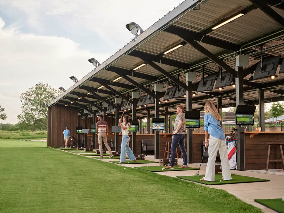 Golf driving range with multiple covered bays, guests practicing, manicured grass, and open field