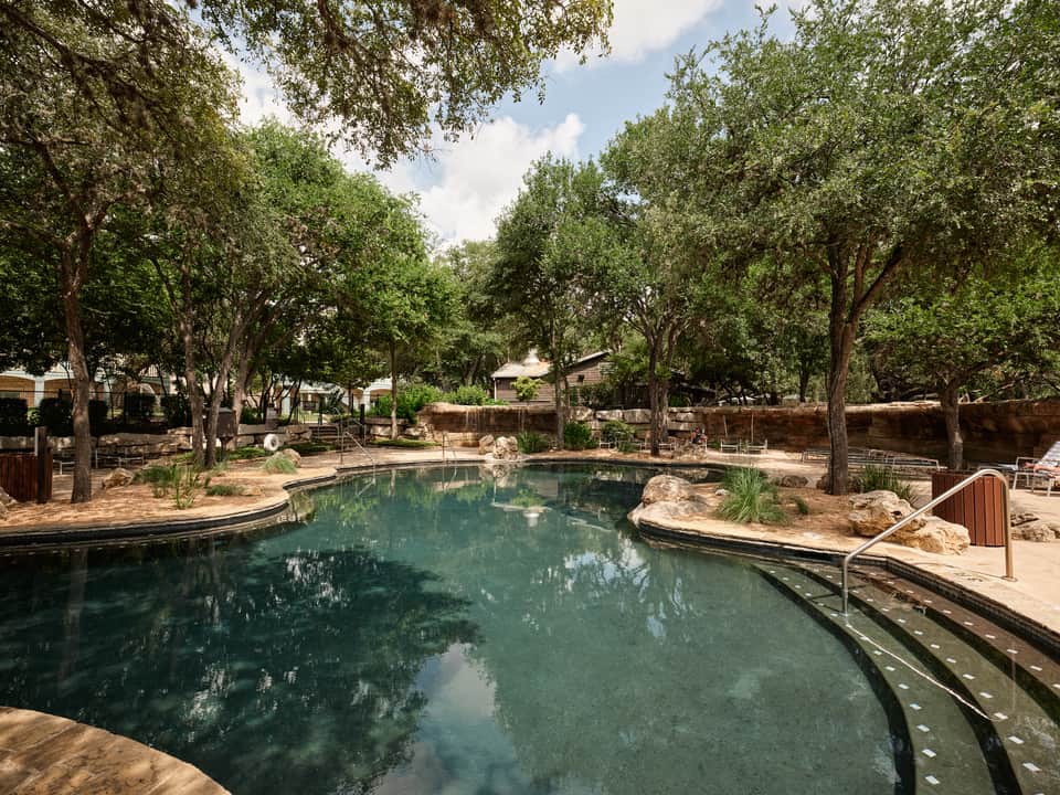 Natural spring-fed swimming pool surrounded by mature trees, stone features, and rustic stone buildings
