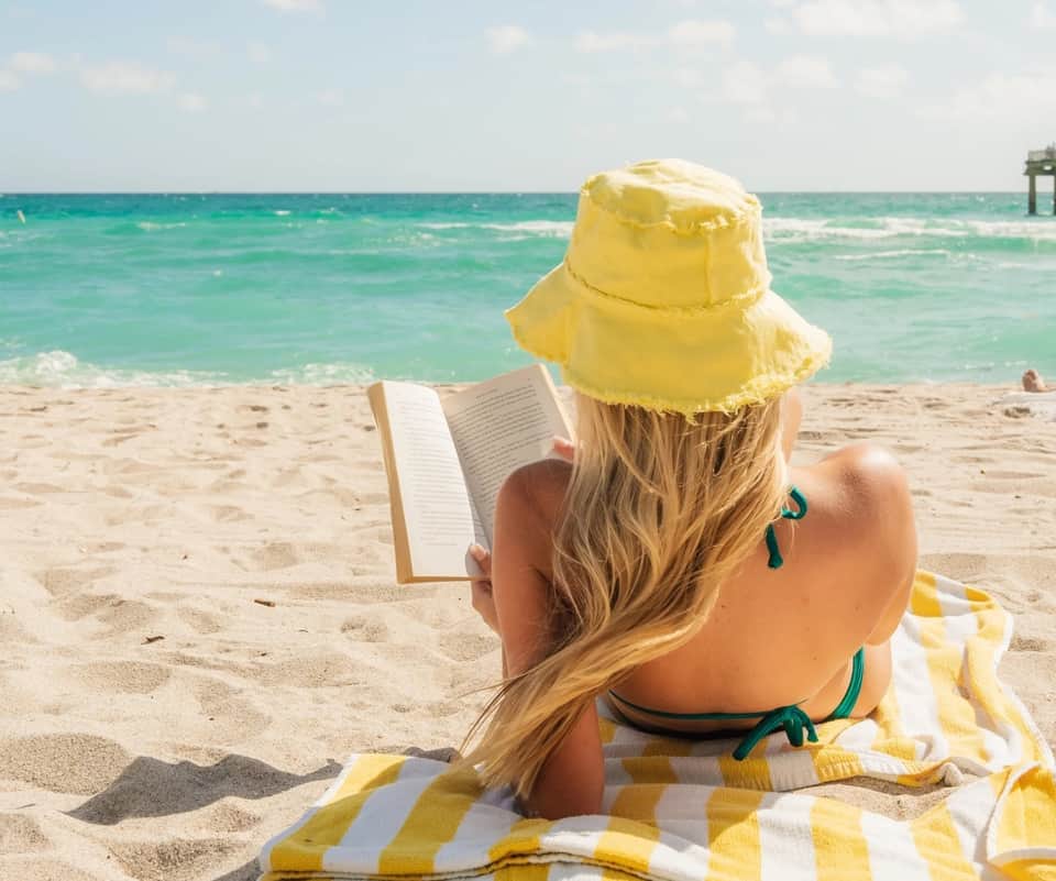 Woman reading a book on beach sand wearing yellow hat and bikini, turquoise ocean and white sand visible