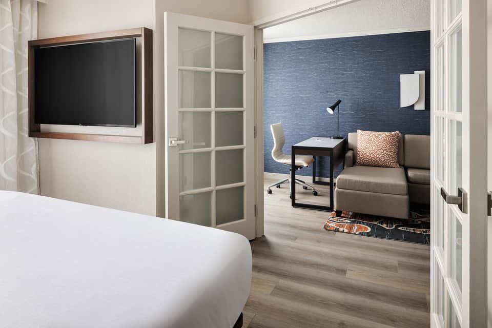 Hotel suite interior showing bedroom with mounted TV and open door to living area with blue accent wall