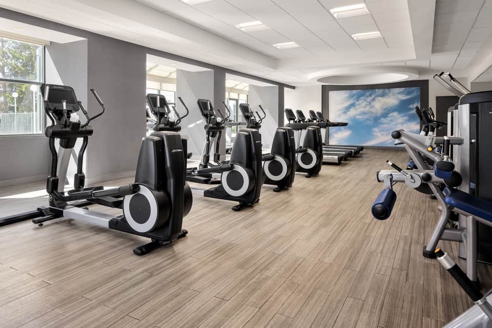 Bright fitness facility featuring rows of stationary bikes, cardio machines, and large screen display