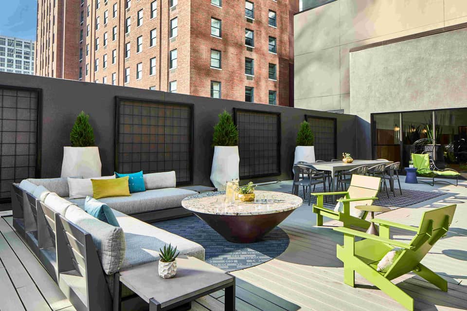 Urban rooftop lounge area with gray sectional seating, round fire pit, lime green chairs, and black privacy screens