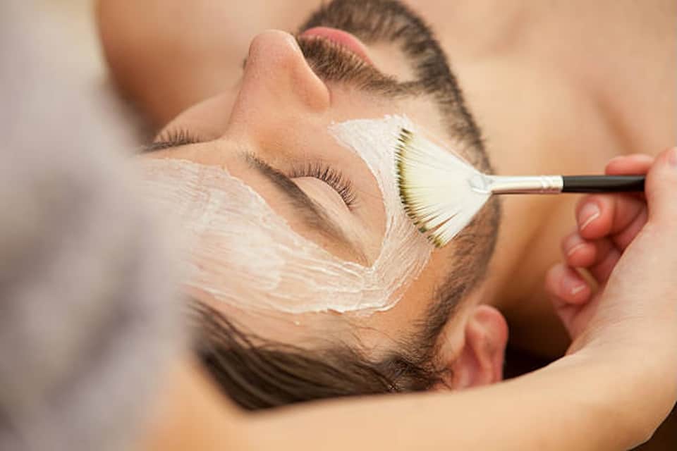 Esthetician applying white face mask to male client with cosmetic brush