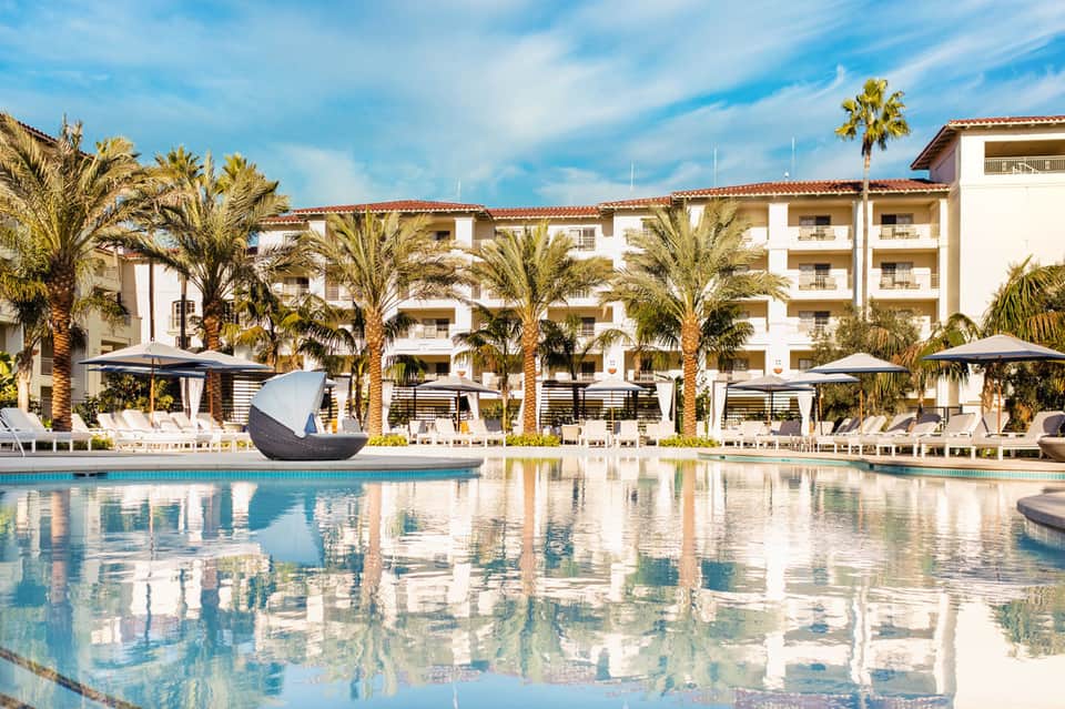 Luxurious resort pool with white loungers, umbrellas, and palm-lined white building complex