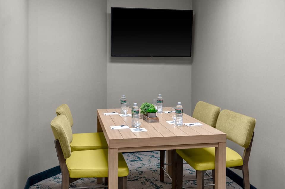 Small meeting room with wooden table, yellow-green chairs, wall-mounted TV, and water bottles