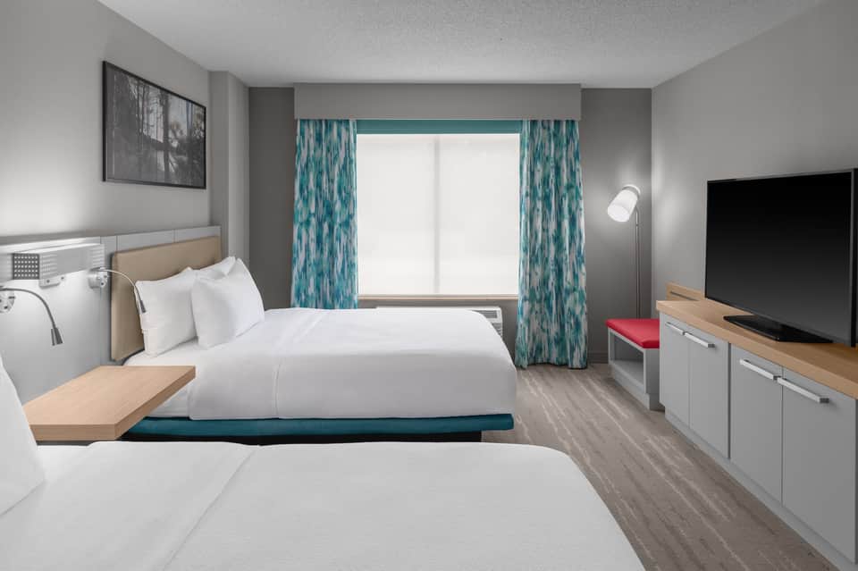 Hotel guest room with two queen beds, teal patterned curtains, TV, and modern furnishings