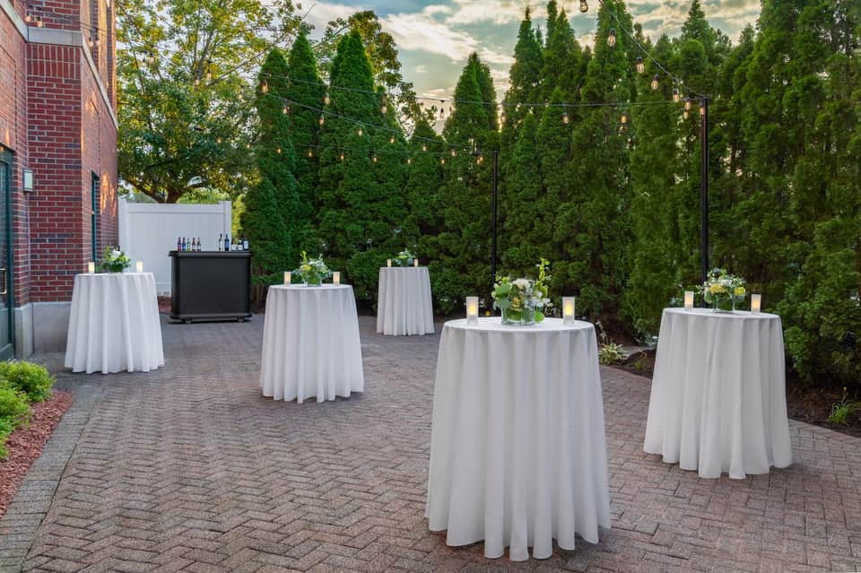 Outdoor cocktail reception with high-top tables, white linens, bar, and green hedge backdrop.
