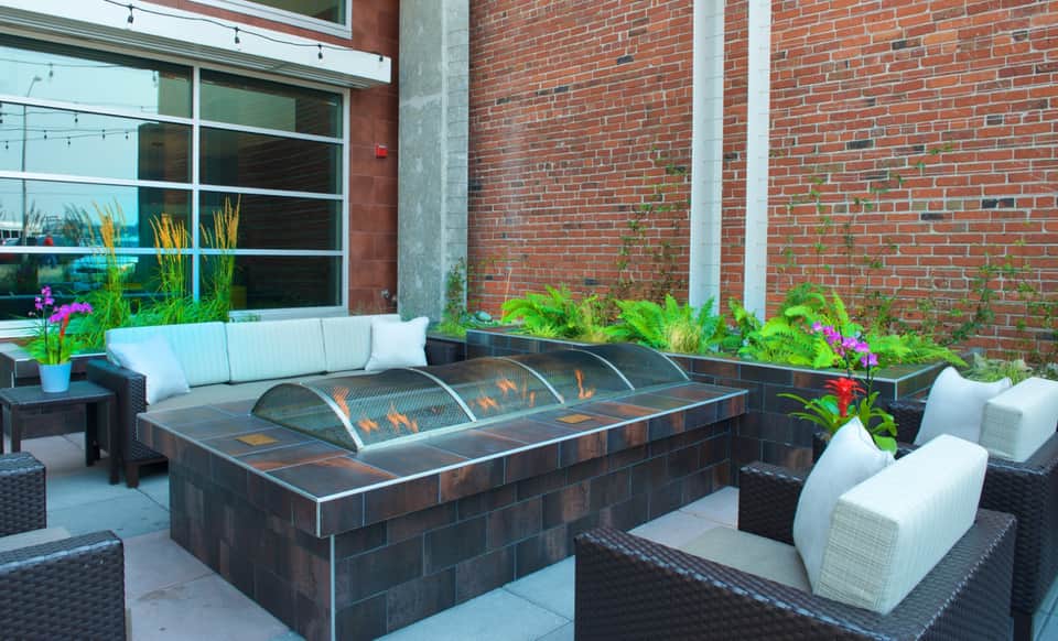 Modern outdoor patio with dark fire pit, white cushioned lounge chairs, and brick building backdrop with purple flowers