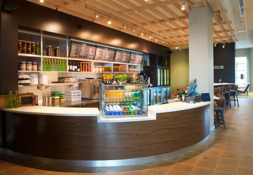 Modern hotel café with curved dark wood counter, beverage refrigerator, menu boards, shelving, and wooden ceiling with track lighting