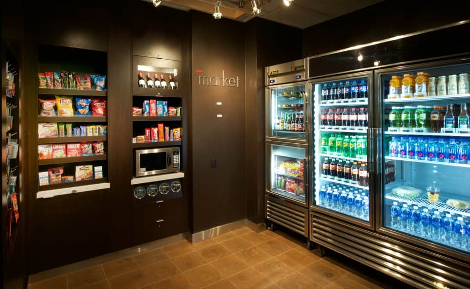 Hotel market nook with vending machines stocked with beverages and snacks, microwave, dark wood shelving, and tile flooring