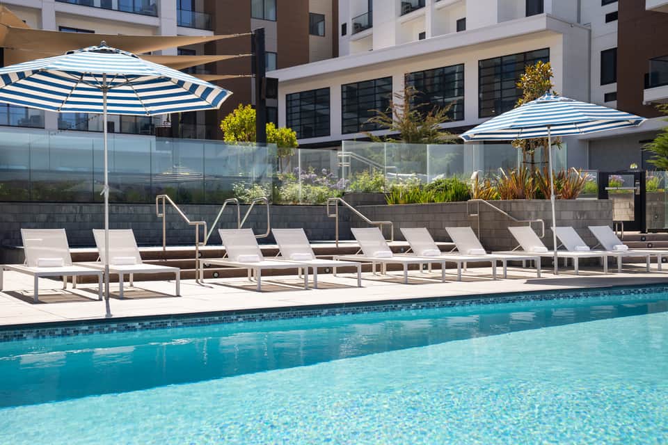 Resort pool deck with lounge chairs, blue striped umbrellas, glass railing, and modern building.