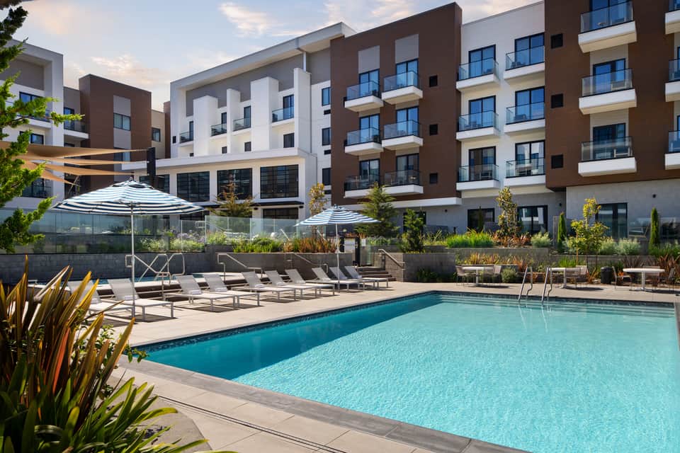Modern apartment complex with turquoise pool, lounge chairs, striped umbrellas, and landscaped terrace