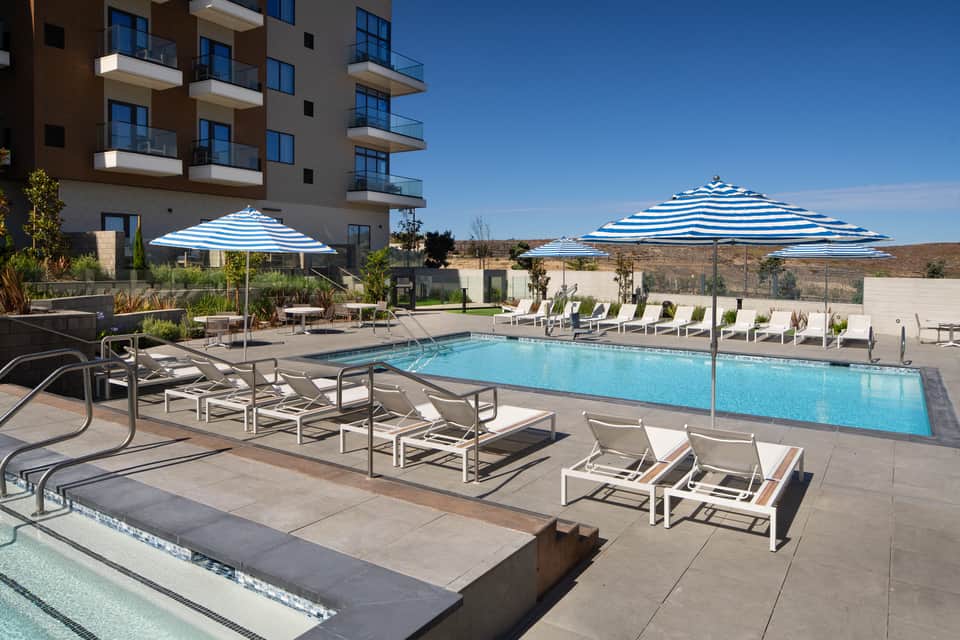Resort pool with striped umbrellas, white lounge chairs, and desert hillside views at daytime