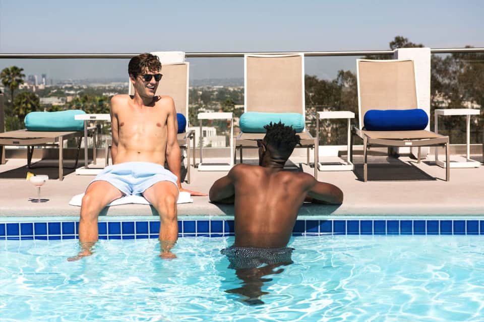 Two men relaxing in shallow resort pool with lounge chairs and city skyline views in background