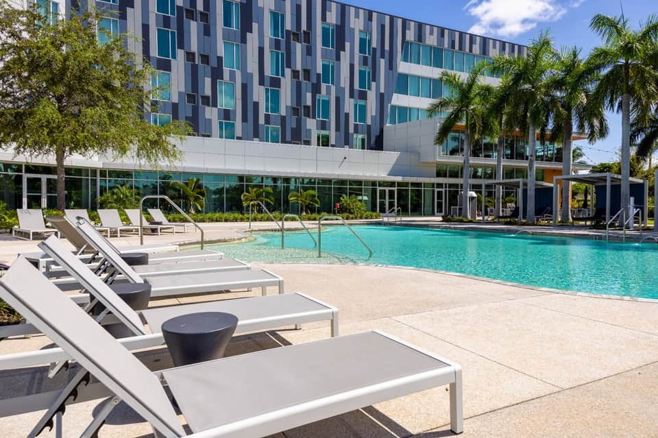 Modern resort pool with turquoise water, lounge chairs, and contemporary high-rise building with palm trees